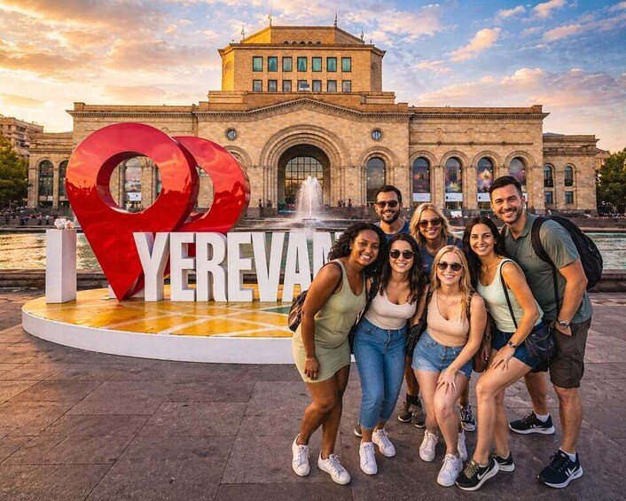 A group of people posing in front of a large red heart-shaped sign with the text 'I YEREVA' and a historic building in the background. 3