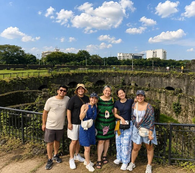 A group of people standing in front of a historic stone structure with greenery and modern buildings in the background. 2