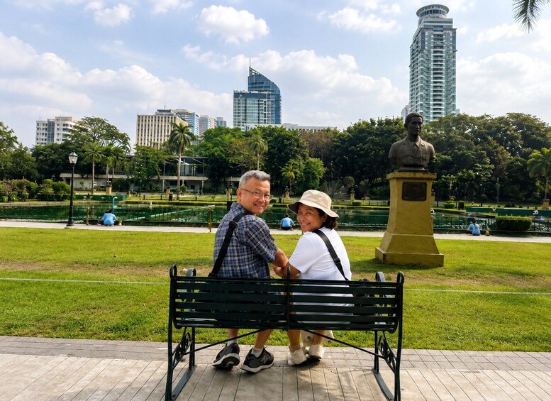 A couple sitting on a bench in a park with a statue and modern buildings in the background. 5