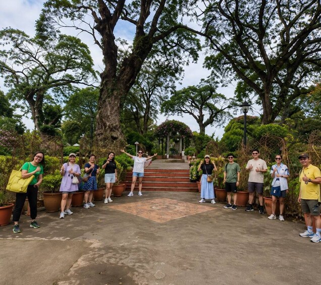 A group of people standing on a concrete path surrounded by large trees and potted plants. 3