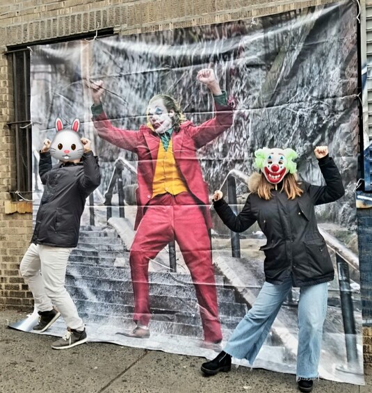 Three people in costumes with bunny and clown masks, standing in front of a large poster. 5