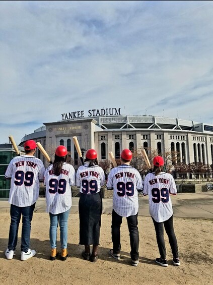 Five people wearing New York Yankees jerseys with the number 99, standing in front of Yankee Stadium. 1