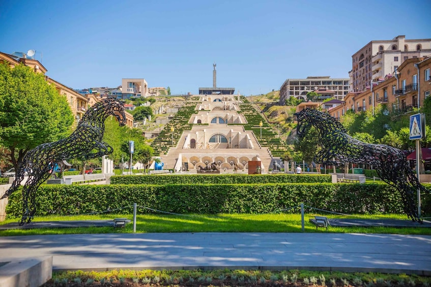 A park with two metal horse sculptures, a central monument, and surrounding buildings. 4