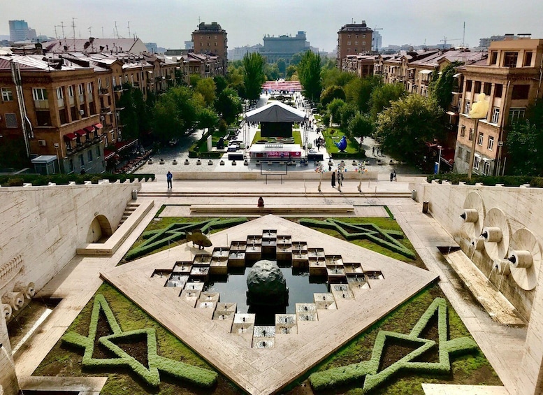 A public square with geometric landscaping, a central fountain, and surrounding buildings. 2