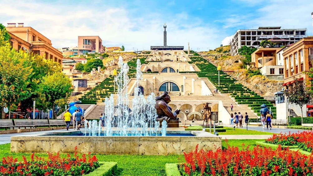 A fountain with water jets, surrounded by red flowers and greenery, with buildings and a statue in the background. 1