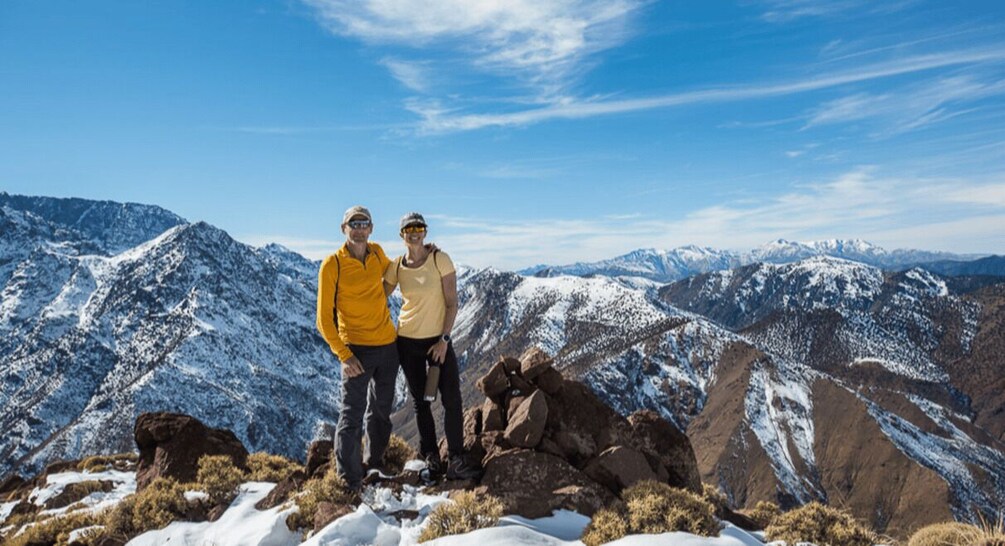 Two people standing on a snowy mountain peak with a vast mountain range in the background. 1