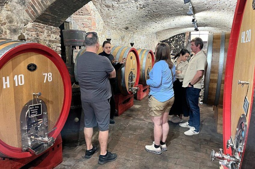 A group of people in a cellar with wooden barrels, some barrels have numbers on them. 4