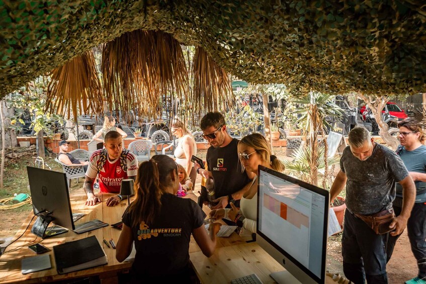 A group of people gathered around a wooden table with laptops and a large monitor, under a thatched roof structure. 4
