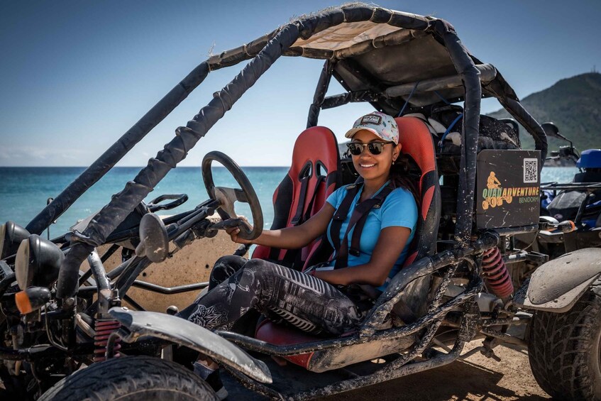A person driving a dune buggy on a beach with a clear sky and ocean in the background. 3