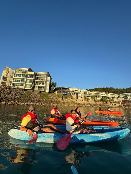 Personas haciendo kayak en un río tranquilo, con casas y una costa rocosa al fondo. 1