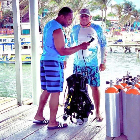 Two people standing on a dock, one holding a device and the other with scuba gear and oxygen tanks nearby. 2