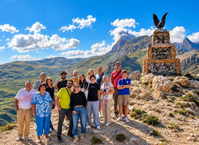 A group of people standing on a rocky outcrop with a mountain range in the background and a stone monument with an eagle statue. 2