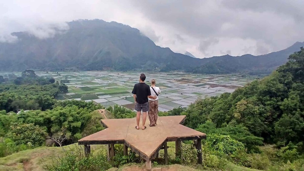 Two people standing on a wooden platform overlooking a terraced rice field with mountains in the background. 1