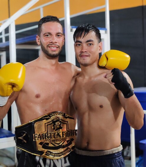 Two men holding a championship belt, standing in a gym with boxing equipment. 5