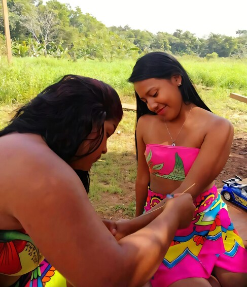 Two people sitting outdoors, one applying a substance to the other's arm. 1