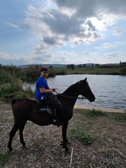 Una persona monta a caballo junto a un río tranquilo con orillas cubiertas de hierba y un cielo nublado. 2