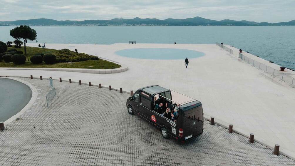 A black van with passengers, parked on a paved area near a waterfront. 1