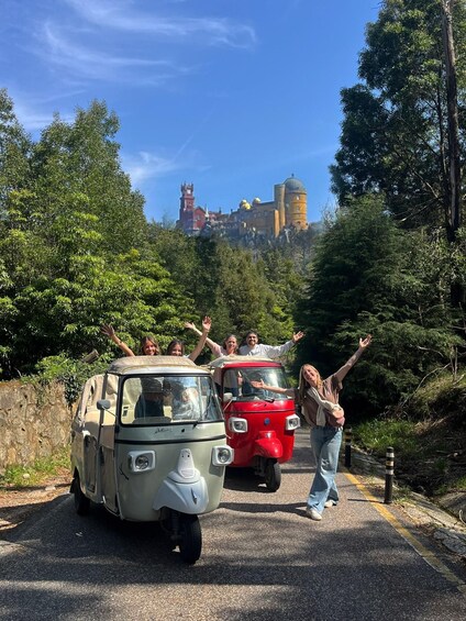 A vintage car and a red car parked on a road with people waving, surrounded by greenery and a castle in the background. 2
