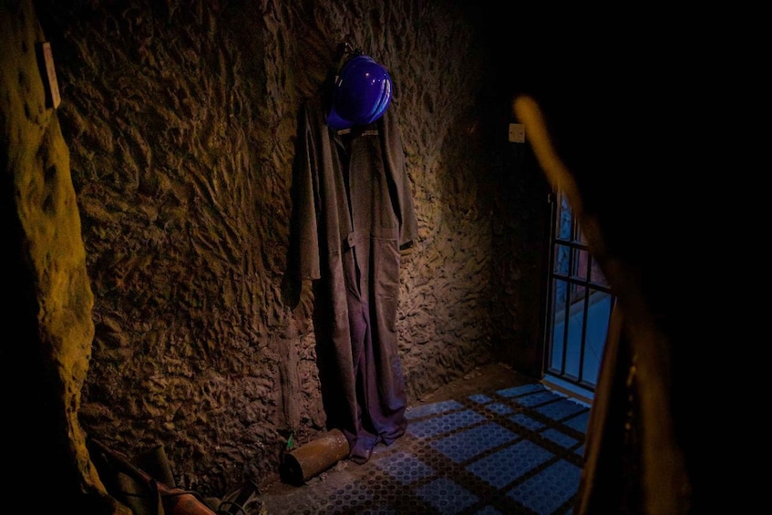 A dimly lit room with a rough stone wall, a blue helmet hanging on the wall, and a window with blue-tinted glass. 3