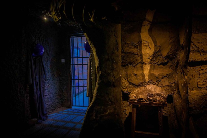 A dimly lit cave with rough stone walls, a wooden table, and a barred window. 5