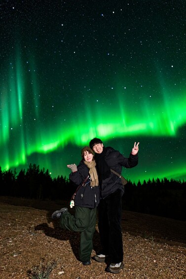 Two people posing under the Northern Lights with a forest in the background. 2