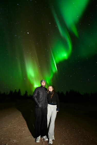 Two people standing on a dirt path under a night sky with green aurora borealis. 3