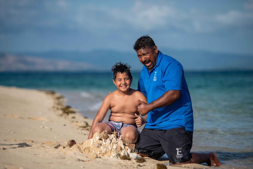 A man in a blue shirt and black shorts kneeling on a sandy beach, giving a thumbs-up to a child sitting nearby. 2