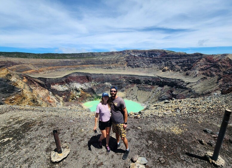 A couple standing on a rocky terrain with a large crater and greenish water body in the background. 1