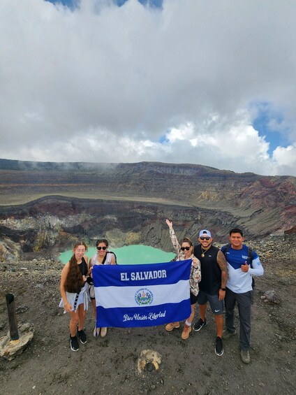 A group of people holding a flag with 'EL SALVADOR' written on it, standing on a rocky terrain with a mountainous landscape in the background. 3