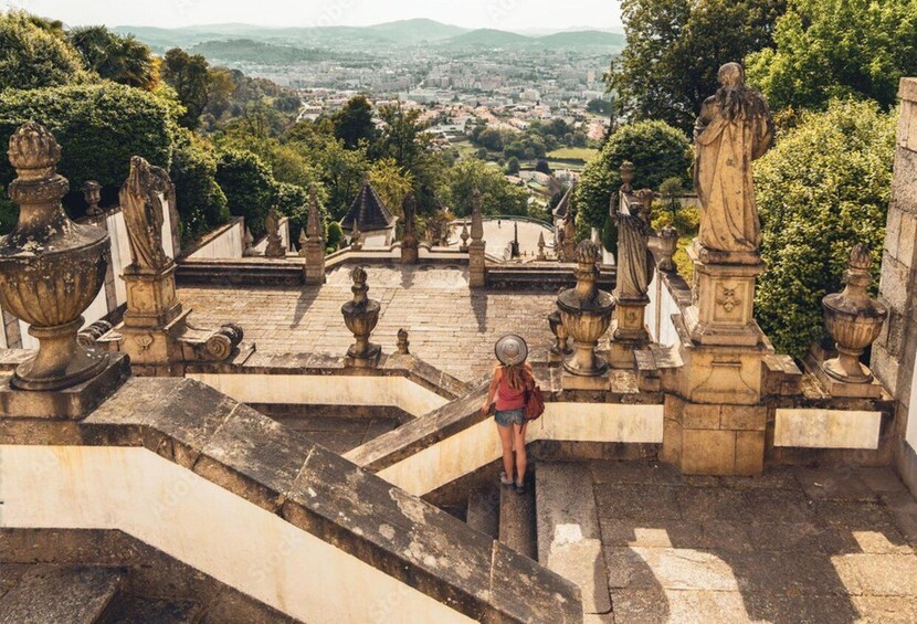 A person standing on a stone staircase with ornate balustrades, overlooking a cityscape with greenery and distant hills. 1