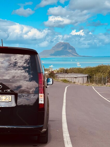 A black car parked on a road with a clear sky, a mountain, and the ocean in the background. 5