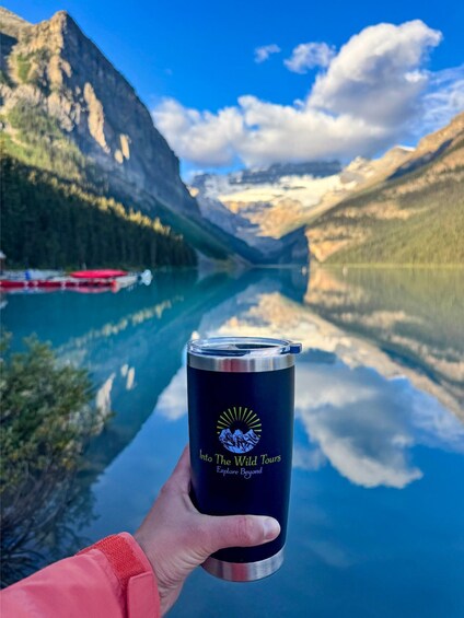 A hand holding a dark blue tumbler with a mountain and sun logo, in front of a mountain lake with a boat in the background. 5