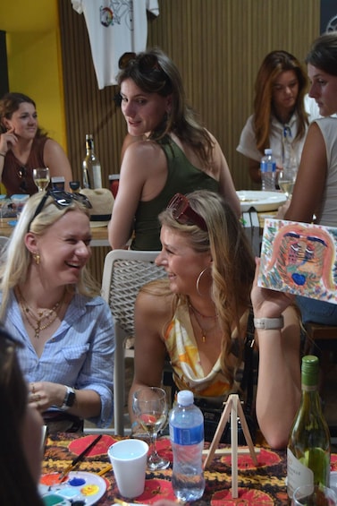 A group of people sitting at a table with a painting, water bottles, and wine glasses. 2