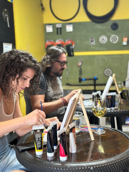 Two people painting on easels with art supplies on a table. 1