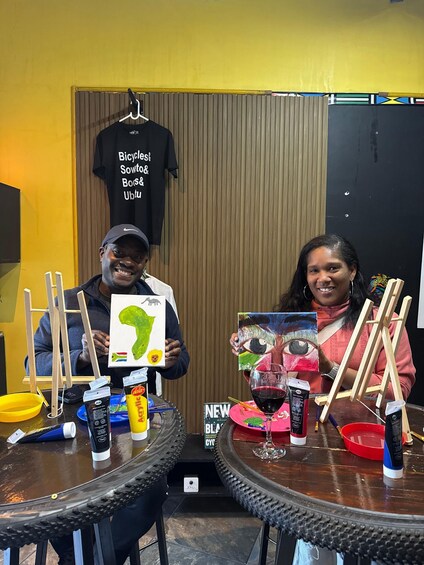 Two people at a table with art supplies, holding paintings, and a t-shirt in the background. 2