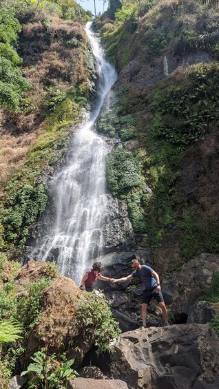 A waterfall in a lush, green valley with two people holding hands at the base. 1