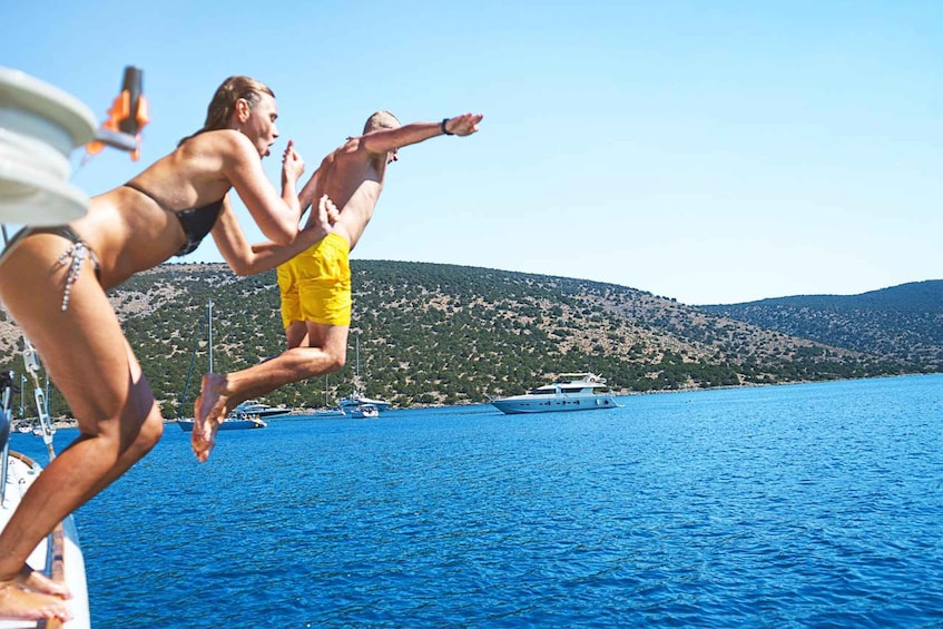 People jumping off a boat into the sea with a yacht and hills in the background. 2