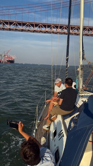 A sailboat with three people on deck, a bridge in the background, and a cargo ship on the water. 5