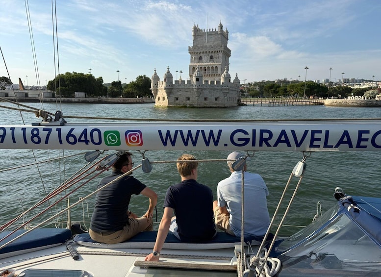 A sailboat with three people, a historic tower, and a river. 3
