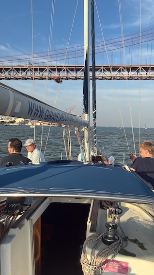 A sailboat with a mast, rigging, and a person holding a device, with a bridge and sailboats in the background. 2