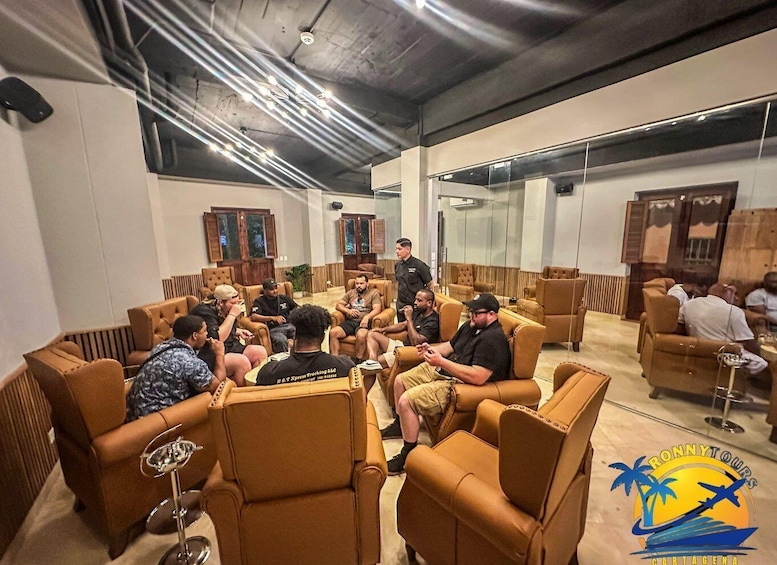 A group of people seated in a lounge area with brown leather chairs, a glass table, and a logo for 'Ronny Tours' on the wall. 5