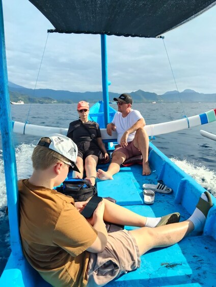 Three people on a boat with a blue interior, a mountainous landscape in the background, and a clear sky. 3