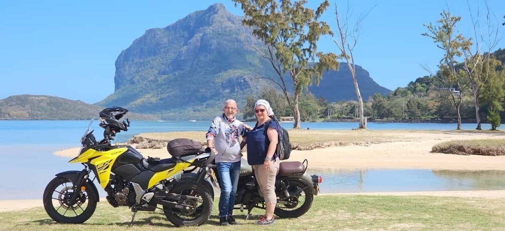 Two people standing next to a yellow motorcycle on a beach with a mountain in the background. 1