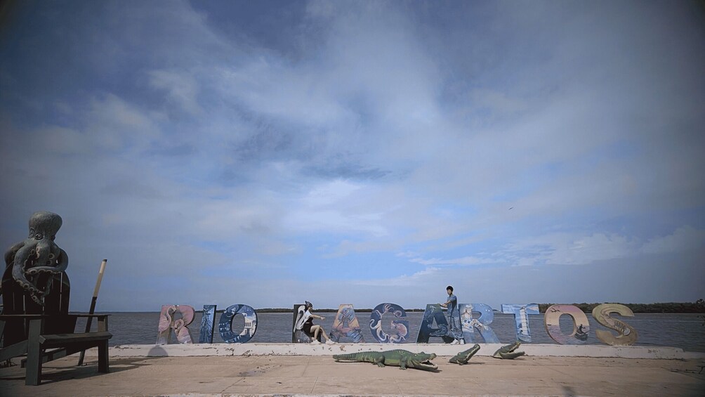A beach with the word "RIO" spelled out in large letters, a statue of an octopus, and a crocodile lying on the sand. 1