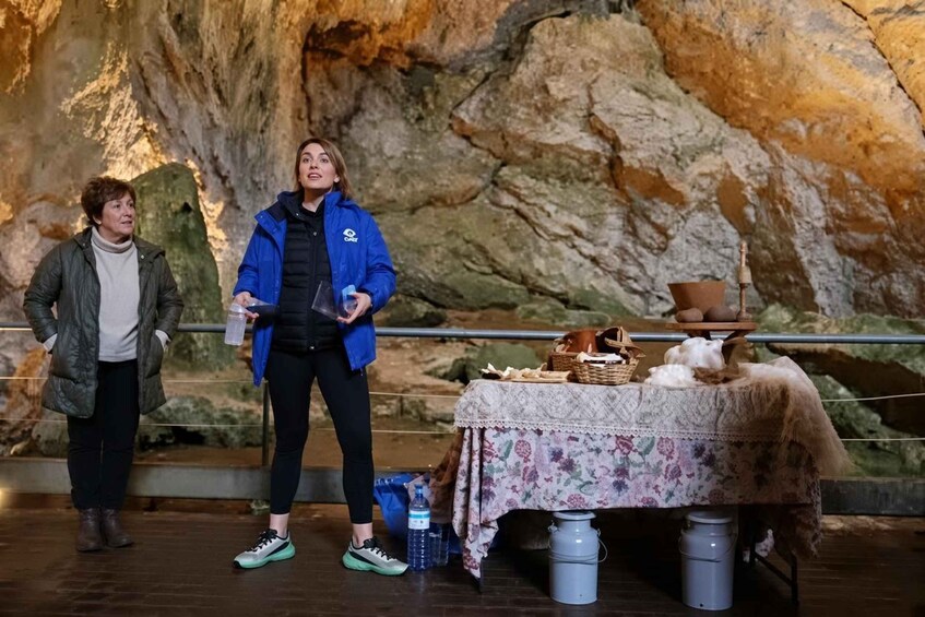 Dos personas en una cueva, con una mesa cubierta por un mantel floral y varios objetos encima. 5