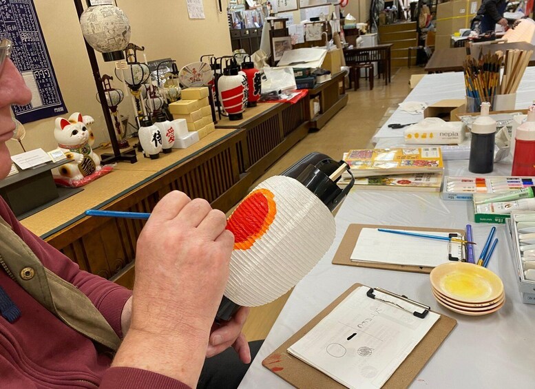 A person holding a white paper lantern with orange and red patterns, surrounded by art supplies and a clipboard with a drawing. 2