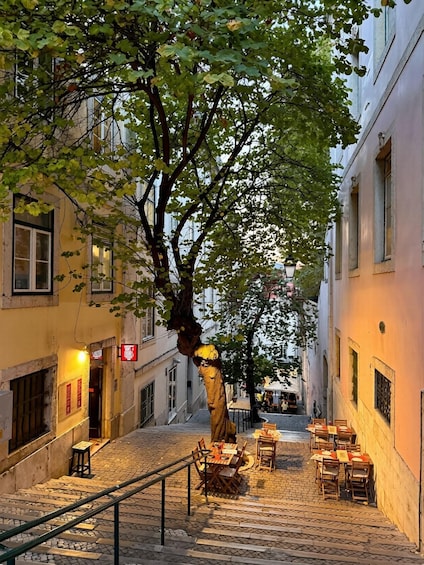 A cobblestone street lined with buildings, a tree with yellow leaves, outdoor seating, and a staircase. 4