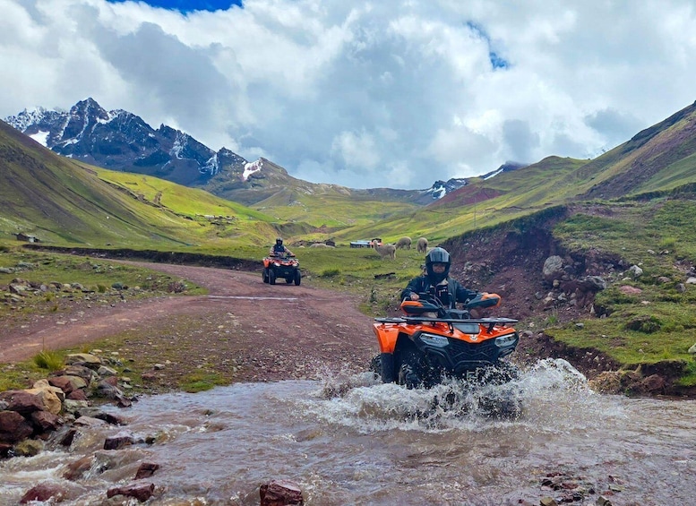 Two riders on ATVs on a dirt road with a stream crossing, surrounded by green hills and mountains. 1