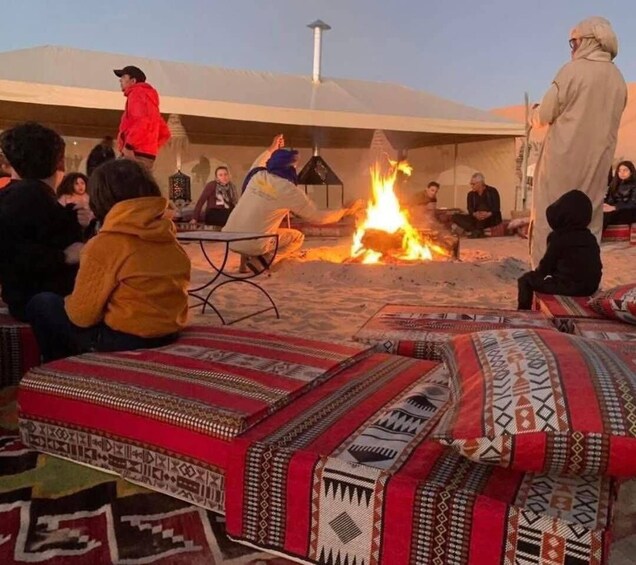 A group of people sitting on patterned rugs around a campfire in a desert setting. 1
