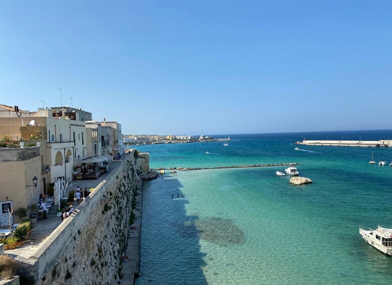 A coastal town with a clear blue sea, boats, and a stone wall along the waterfront. 1